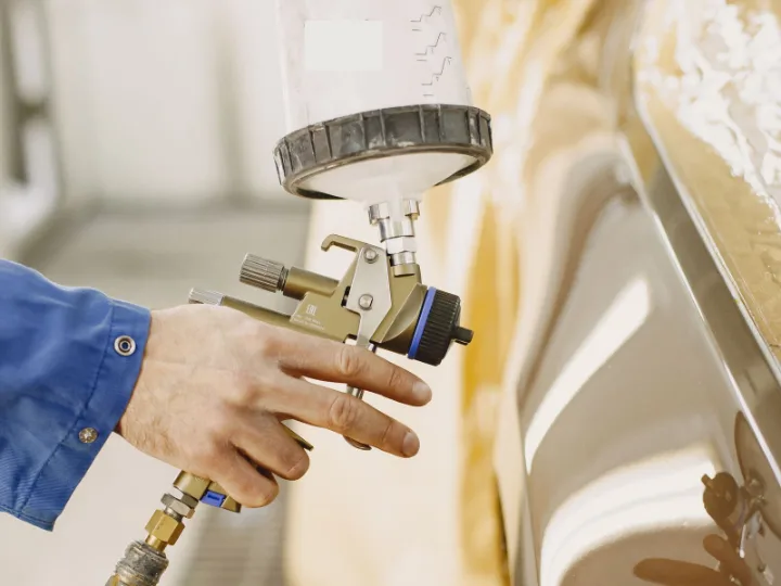 Hand met een spuitpistool naast een afgeplakte auto in de spuitcabine
