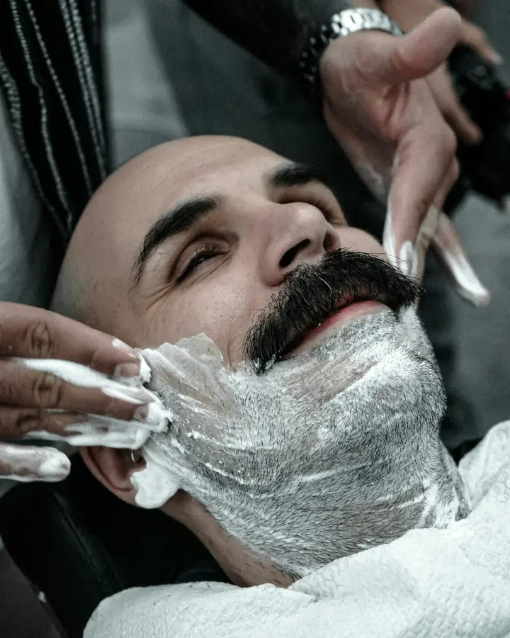 Een klant met een volle snor ligt achterover terwijl de barbier scheerschuim op zijn wangen aanbrengt