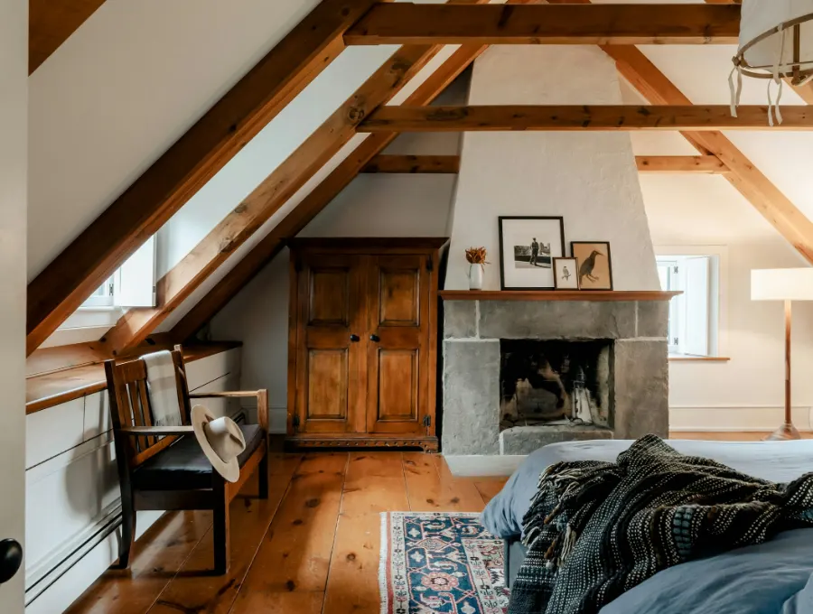 Zolderkamer onder houten balken met een open haard, een fauteuil en een groot bed