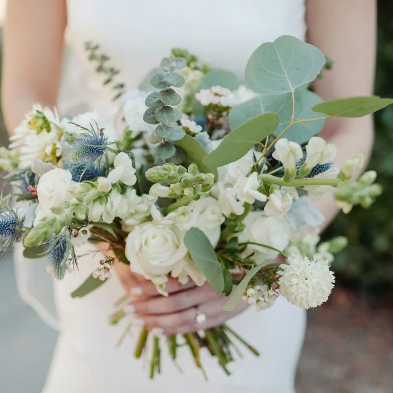 Een bruid houdt een los boeket van witte rozen, blauwe distels en eucalyptus vast