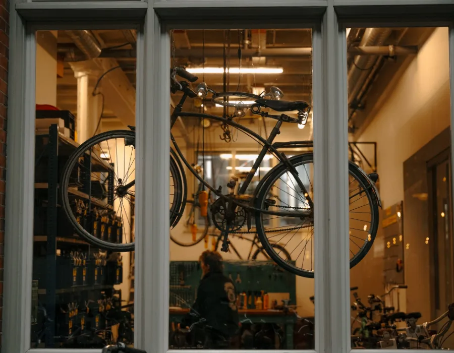 Door het raam van de werkplaats: een fiets hangt aan het plafond, daaronder werkt iemand aan de werkbank