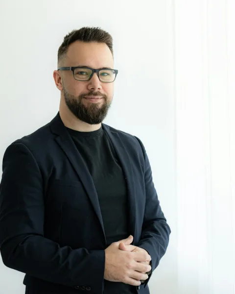 Portret van een man met bril en baard in een donker colbert voor een licht raam