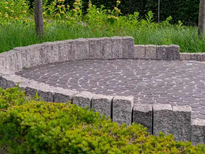Na: rond terras van donkere natuursteen in cirkels, omringd door granieten stapelblokken en lage beplanting