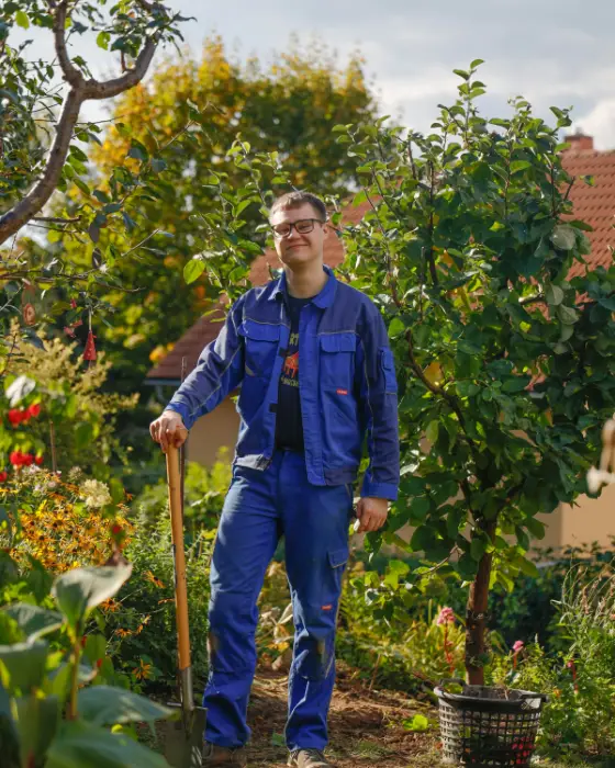 Wietse in een blauwe werkoverall staat met een schop in een bloeiende tuin