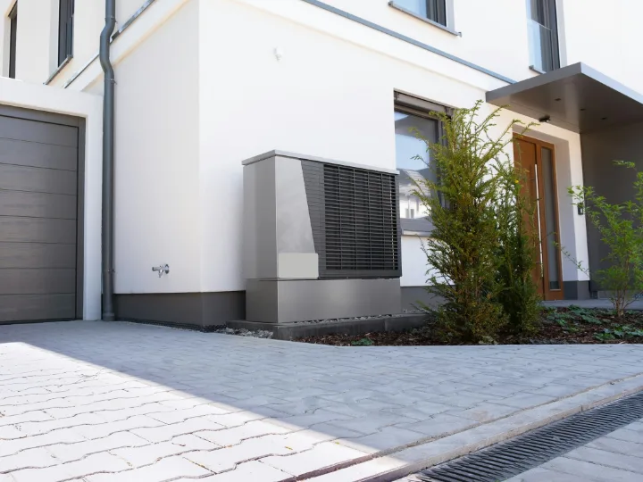 Buitendeel van een warmtepomp naast de voordeur van een wit huis met oprit