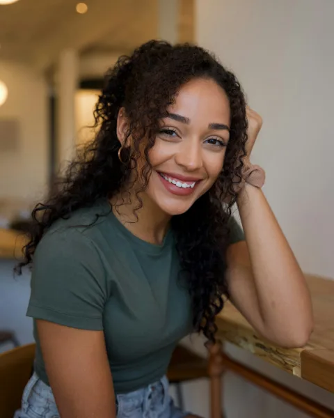 Noor met lang krullend haar, lachend aan een houten tafel
