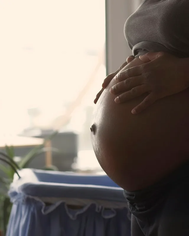 Een zwangere vrouw in een grijs shirt legt haar hand op haar buik bij een licht raam