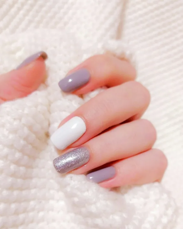 Nagels in mauve en wit met één zilveren glitternagel, tegen een wit gebreid vest
