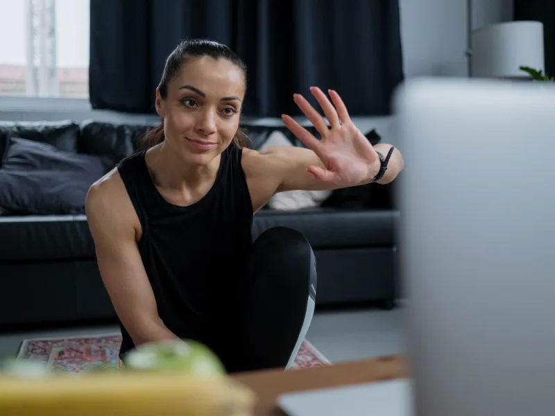 Een vrouw zwaait naar haar laptop terwijl ze thuis op de vloer traint