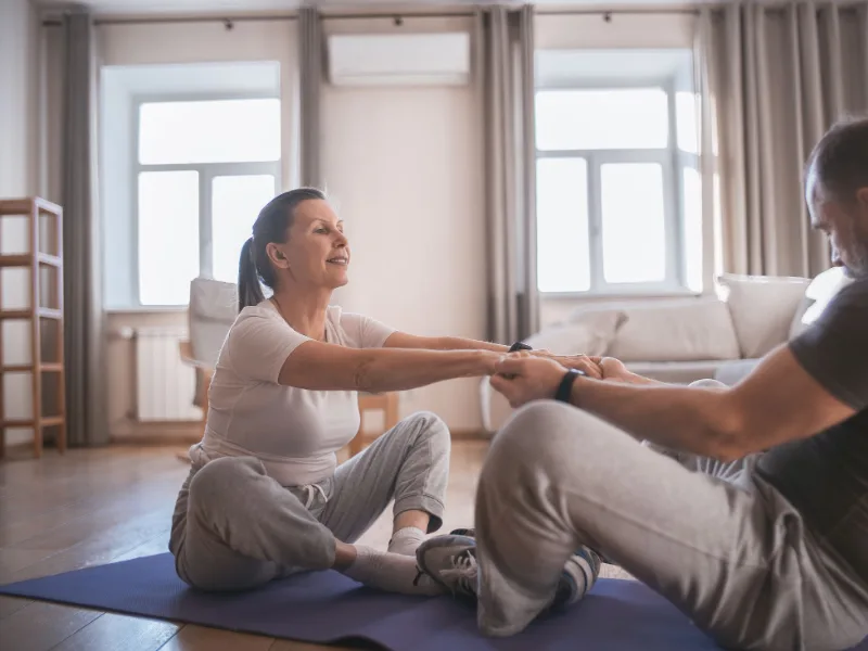 Een trainer en een oudere vrouw doen samen een rekoefening op een mat in de woonkamer