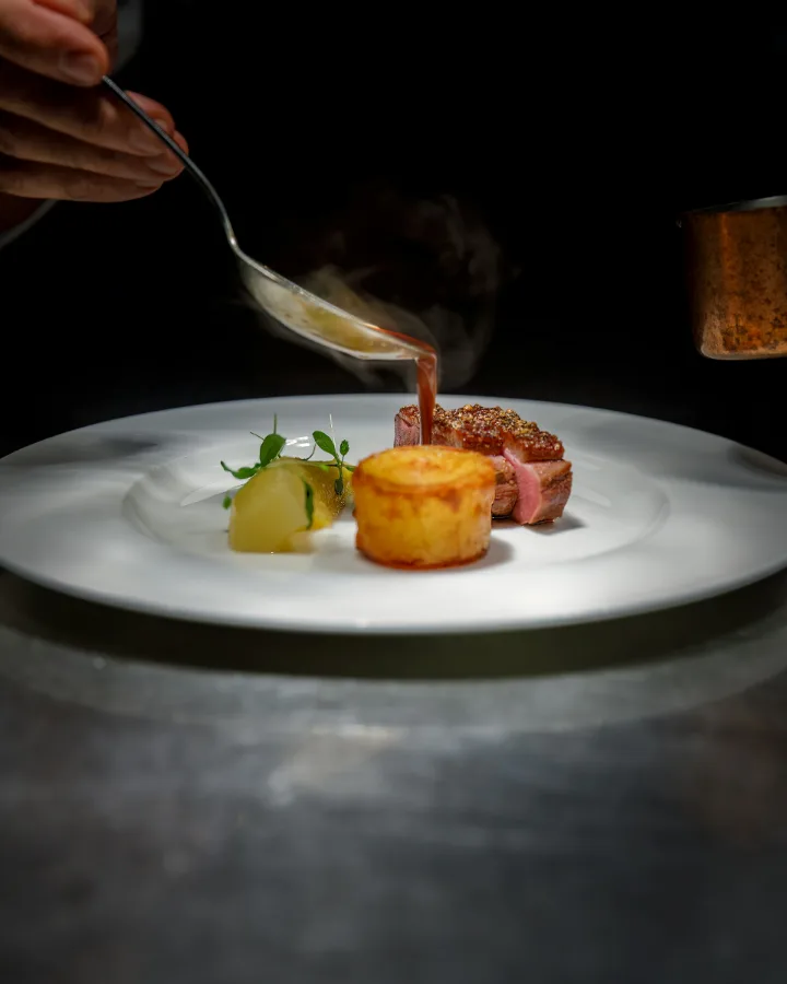 De chef schenkt saus over een bord met vlees, aardappel en groente, in een donkere keuken