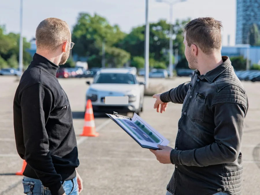 Een examinator met een klembord wijst een leerling op een parkeerterrein met pionnen de weg