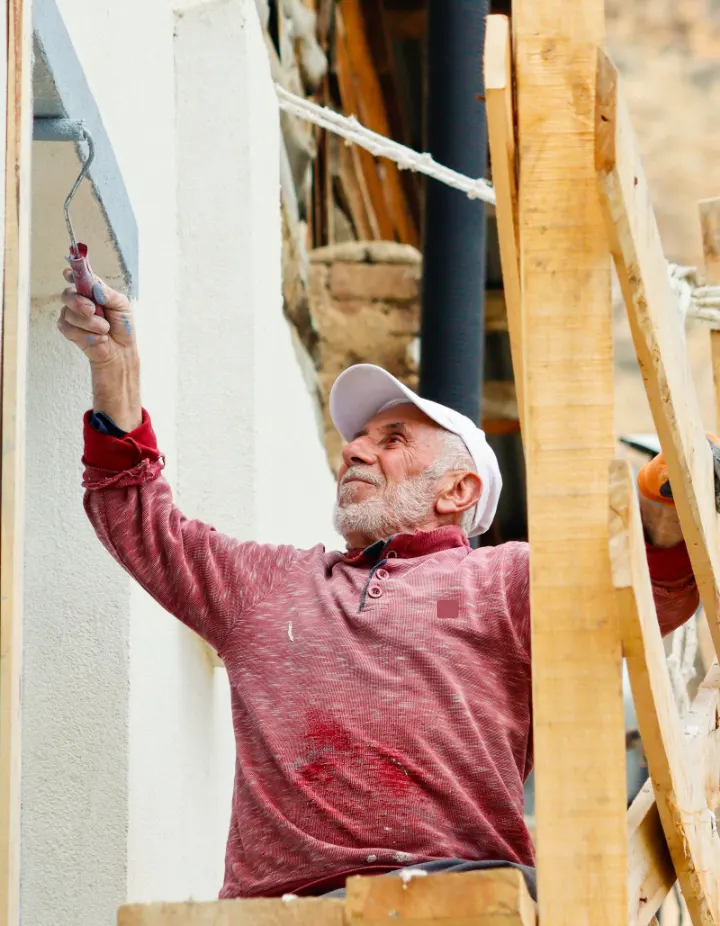 Oudere schilder in een rode trui rolt witte verf op een gevel vanaf een houten steiger