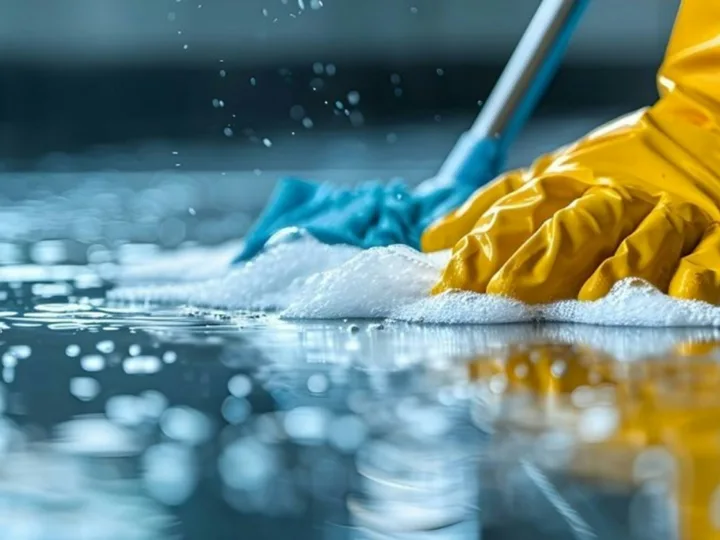 Een hand in een gele handschoen schrobt met een blauwe doek een natte vloer vol sop