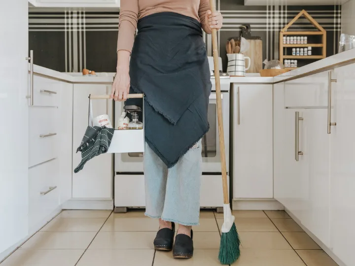 Een vrouw met een schort en een bezem staat in een lichte keuken
