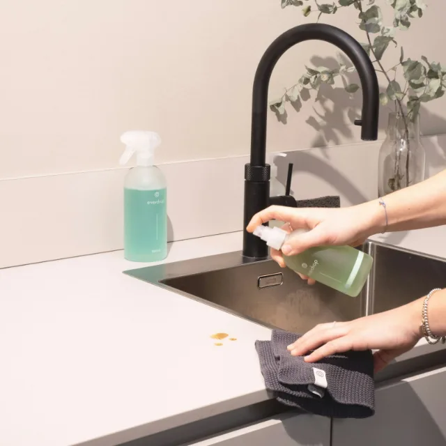 Een hand veegt met een doek een zwarte spoelbak schoon, naast een sprayflacon
