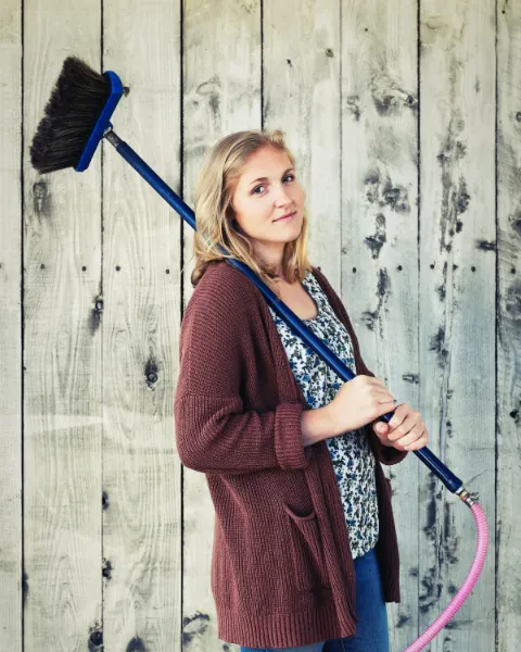 Sytske staat lachend met een bezem over haar schouder voor een houten wand
