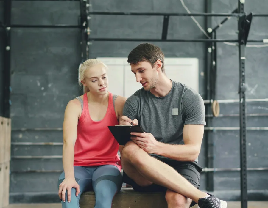 Een trainer schrijft op een tablet terwijl hij met een sporter op een bank in de krachthal zit