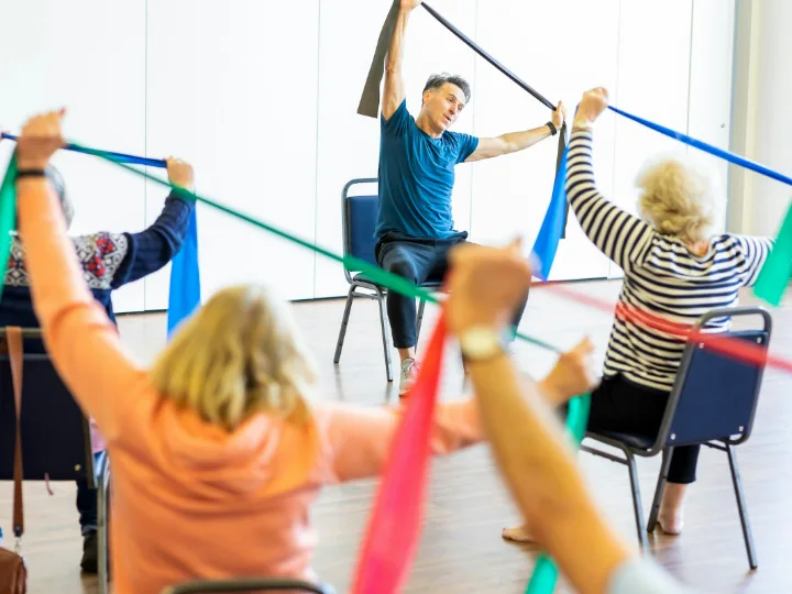 Een groep oudere sporters rekt elastieken banden uit boven het hoofd, met de trainer voorin