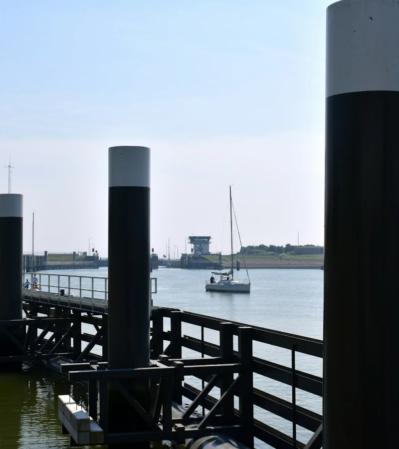 Houten steiger met meerpalen aan het water, met in de verte een zeilboot